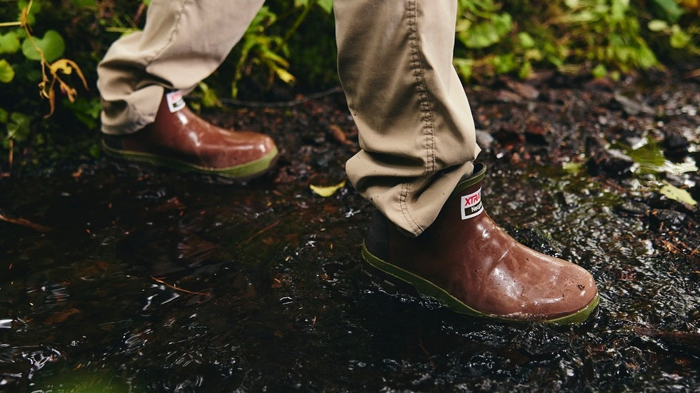 Man wearing Xtratuf waterproof Legacy boots walking through muddy terrain, showcasing slip-resistant soles and rugged fit for wet, tough environments.