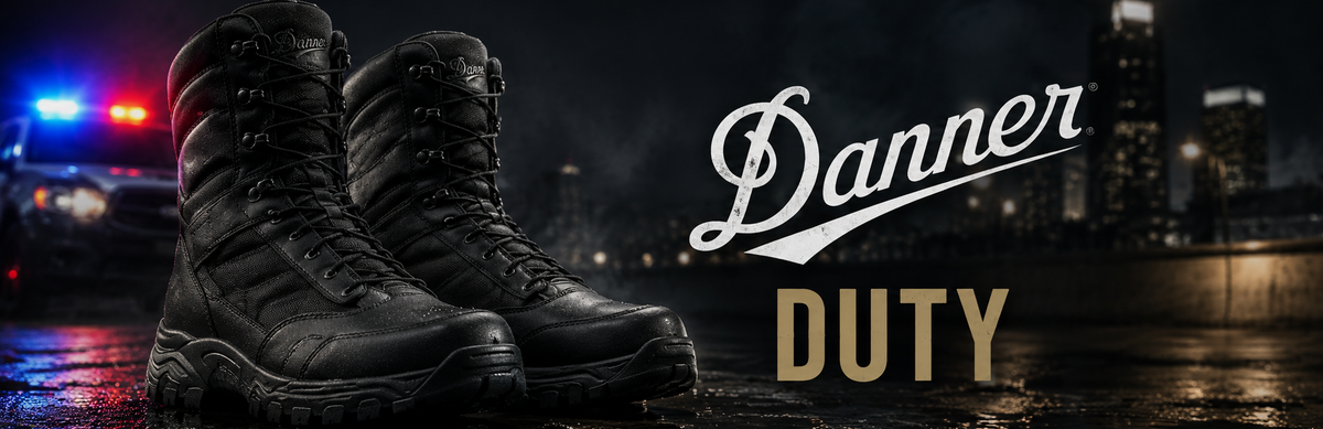 A pair of black Danner Duty tactical boots standing on wet pavement at night, with a blurred city skyline and police lights in the background. The words 'Danner' and 'DUTY' appear on the right side.