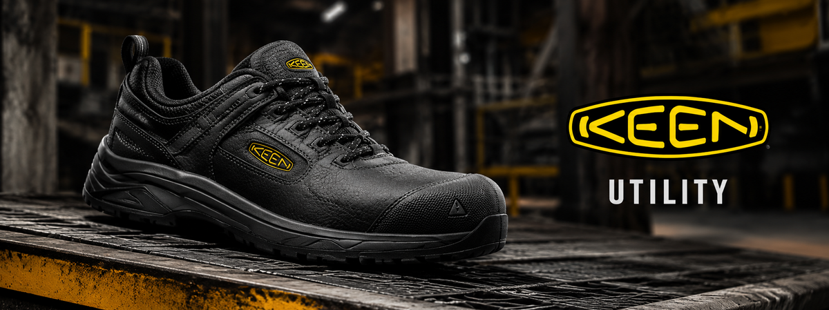 A black Keen Utility work shoe with a textured sole, displayed on a wooden platform in an industrial setting. The Keen logo and the word 'UTILITY' are visible on the right side of the image.